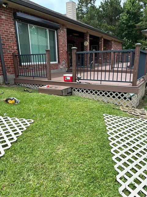 Deck with black railing beside a brick house, lattice skirting panels laid out on the grass